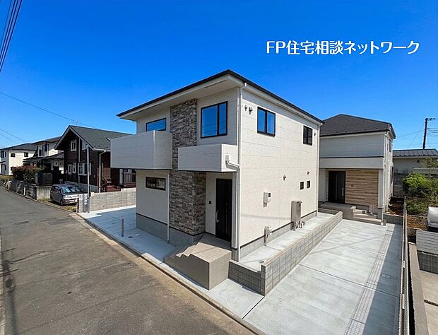 【外観】白い外壁に石目調のアクセントを取り入れた2階建ての住宅です。前面には広いコンクリート敷きの駐車スペースが設けられ、複数台の駐車に対応。落ち着いた雰囲気の住宅街に建っています。