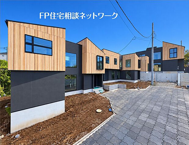 【分譲済街並】 青空を背景に、木目調の外壁とダークグレーのコントラストが印象的なモダンな建物が連なっています。