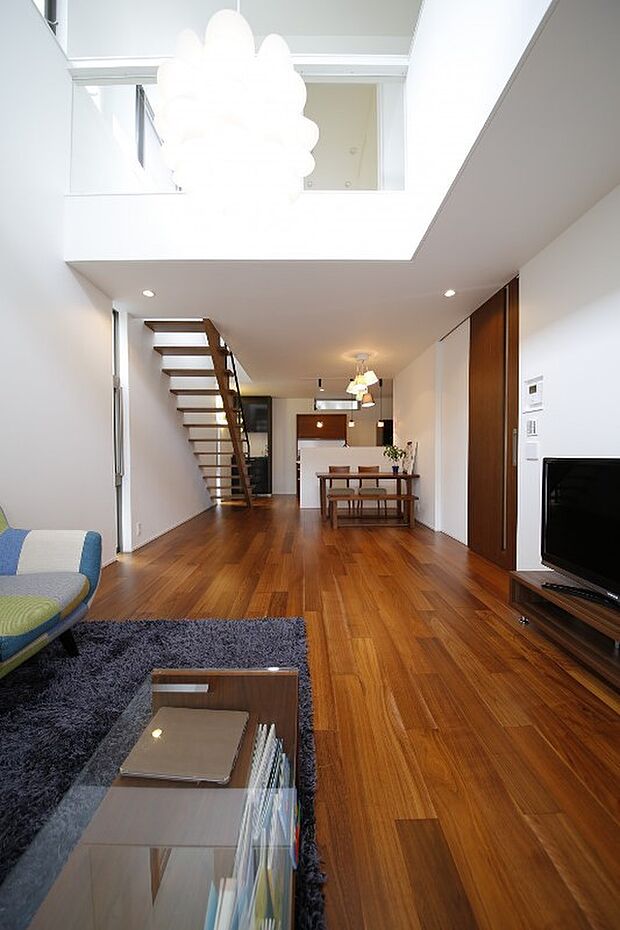 【建物プラン例（内観写真）】大きく張り出したバルコニー、その下のデッキスペースを挟んでリビング。奥まったリビングへは大きい吹き抜けから光を取り込みます。1Fの開口はデッキスペースへの大きな開口以外を絞っているため吹き抜けから落ちてくる光がより強調されます。またバルコニーに面した大きな吹き抜けの窓は格子や反射光により様々な表情を見せます。外部からのプライバシーを守りながらも、光溢れる落ち着いた空間になりました。/当社施工例