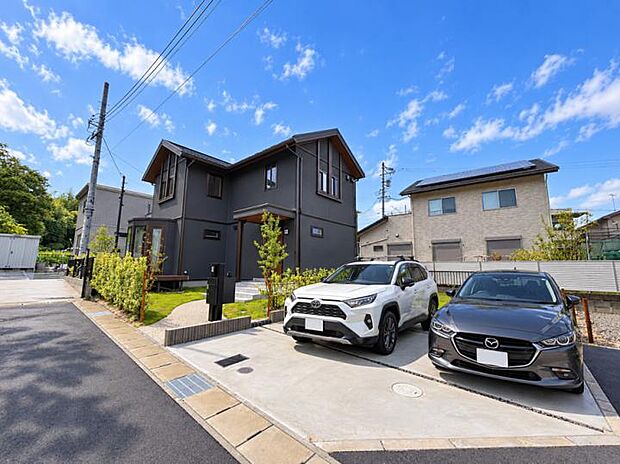 【【B号地】】ゆとりある敷地に車2台をゆったり並列駐車できるスペースを確保。青空に映えるシックな外観と洗練された外構計画が、街並みの中で一際存在感を放ちます。※車はAI合成しています