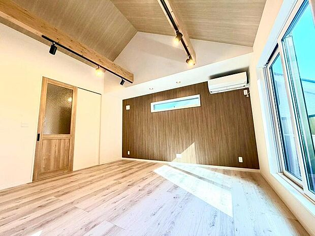 勾配天井と木の梁が開放感を演出するリビング。木目調のアクセントウォールが温かみを添え、家族の笑顔が自然と集まる空間。明るい陽光が降り注ぎ、心地よい毎日を育みます。