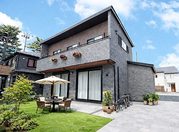 【施工例外観】落ち着いたグレーの外観が街並みに調和するモダン邸宅。広々とした庭とテラス空間が、家族の時間を豊かに彩ります。バルコニーの花々がアクセントとなり、上質さと温もりを感じる住まいです。