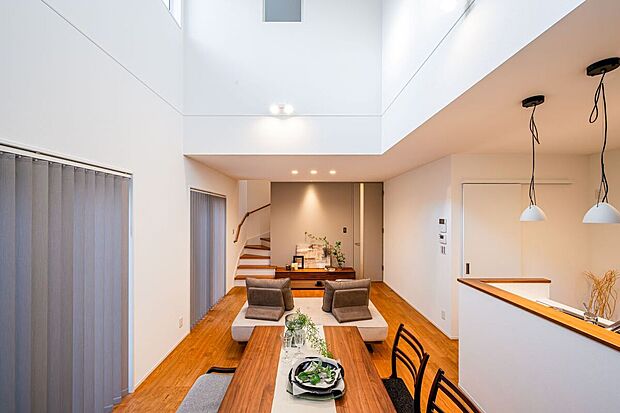 A号棟リビング。「上質な暮らしを叶える、光あふれるLDK」階段が空間のアクセント。白壁と無垢材フローリング、落ち着いた家具でモダンかつ居心地の良いLDK。大きな窓の陽光が家族の団欒を優しく包みます