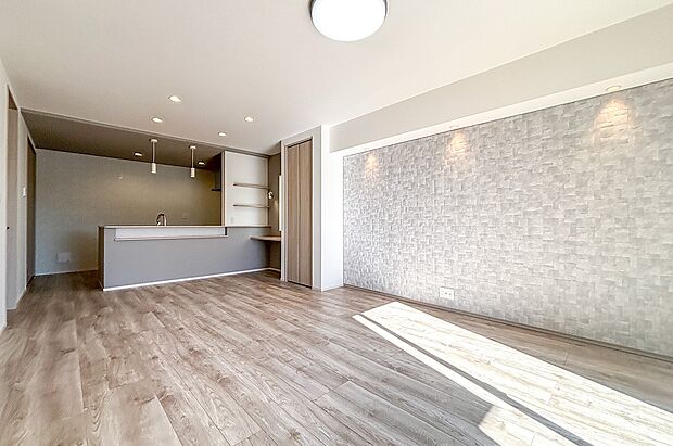 明るく広々としたリビング。木目調のフローリングとアクセント壁が温かみを演出し、ダウンライトが空間をやさしく照らします。キッチンとの繋がりもよく、家族が集まりやすい快適な住まいです。（2号棟）