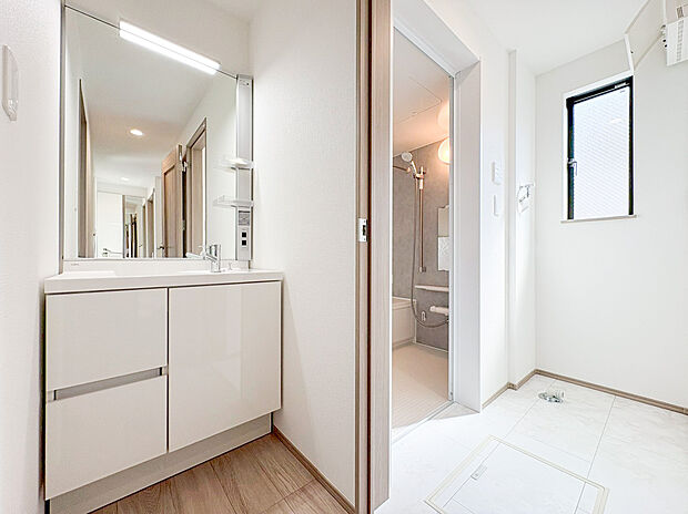【洗面化粧台】白を基調とした清潔感あふれる洗面空間。大型の鏡と豊富な収納を備えた機能的な洗面化粧台は、毎朝の身支度をスムーズにしてくれます。浴室と隣接し、家事動線にも配慮された使い勝手の良い設計です。