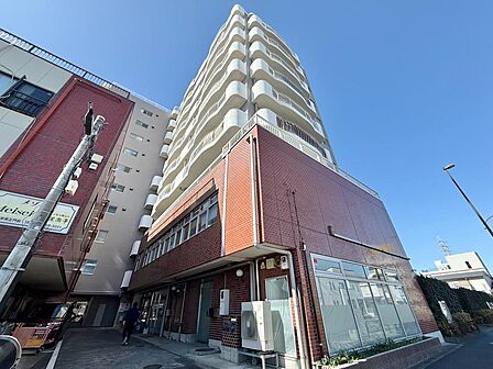 JR武蔵野線「北府中」駅徒歩5分の立地に建つマンション外観です。赤レンガ調の低層部と白いバルコニーが重なるデザインが印象的で、9階建の存在感ある佇まい。駅近ながら落ち着いた住環境が魅力です。