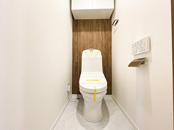 深みのある木目調のアクセントクロスが、ホテルのような落ち着きを醸し出すトイレ空間。毎日を快適に過ごせるリモコン付きの温水洗浄便座を採用しています。上部にはトイレットペーパーのストックに便利な吊戸棚付き