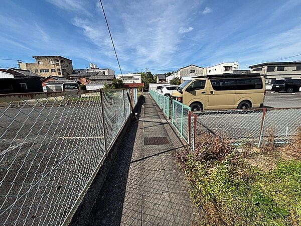 青空が広がる晴れた日の現地写真です。フェンスの向こうには駐車場、その奥には建物が立ち並んでいます。周辺環境をご確認いただけます。