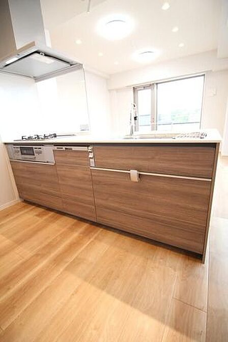 あたたかみのある木目調パネルを採用したシステムキッチンには、3口ガスコンロやグリル、スライド式収納を備えており、天井のダウンライトや木目調フローリングとともに統一感のある空間に仕上がっています。