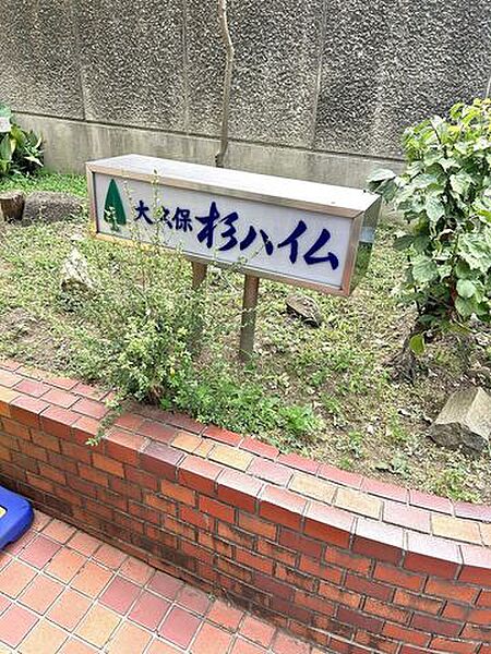 赤茶色のレンガ調タイルが敷かれたアプローチの脇に、植栽が配された花壇が設けられており、その中にはステンレスフレームで囲まれたスタイリッシュなデザインのマンション銘板が設置されています。