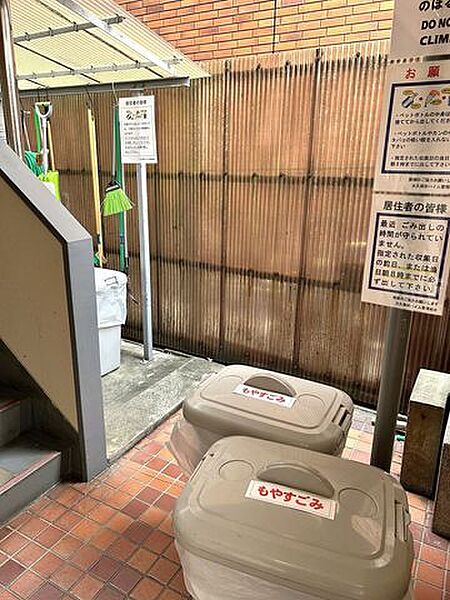 敷地内の共有スペースには「もやすごみ」と書かれた蓋付きのゴミ箱が設置されており、壁面にはゴミ出しルールに関する案内板が掲示されているほか、ほうきなどの清掃用具が整理されて置かれています。
