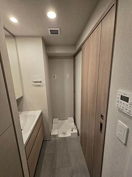 木目調デザインの洗面化粧台と奥に洗濯機パンが配置された洗面室で、天井にはダウンライトが設置されており、統一感のあるナチュラルな木目調の建具と落ち着いたグレー調のフロアで構成されています。