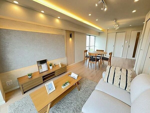折上天井に施された間接照明や多数のダウンライト、スポットライトに加え、グレーのアクセントクロスを照らす間接照明など、多彩な照明設備が整えられた木目調フローリングの室内空間です。
