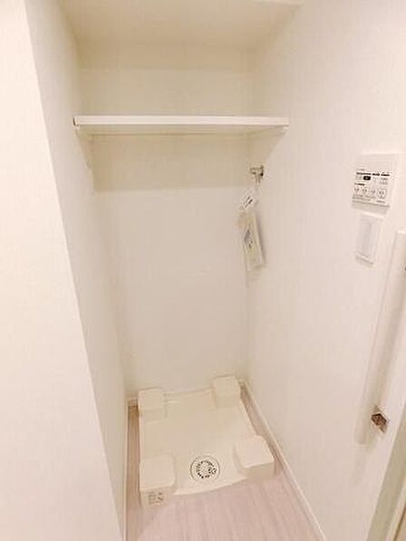 室内には専用の洗濯機パンが設置されており、上部には洗剤や備品の整理に役立つ固定棚が備え付けられております。全体が白で統一された清潔感のある空間となっております。