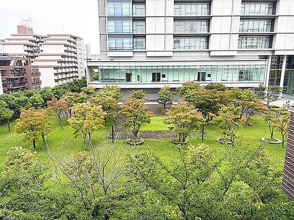 バルコニーからは多くの樹木や芝生が整えられた緑地スペースが見え、その奥にはガラス張りの大型建築物や周辺の建物群が確認できるロケーションとなっています。
