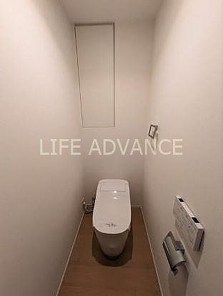 白を基調としたシンプルなトイレには、温水洗浄便座や壁掛け式のリモコン、ペーパーホルダーが備え付けられており、正面奥の壁面には扉付きの埋め込み収納棚が設置されております。
