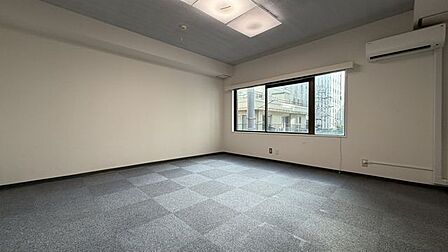 床面にグレーのタイルカーペットを採用した室内には、エアコンやスクエア型の天井照明が備わっており、壁面にはブラインド付きの大きな引き違い窓が設置された清潔感のある空間となっています。