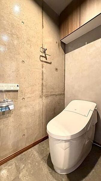 コンクリート打ちっぱなしのデザイン壁が印象的なトイレには、温水洗浄便座機能付きのトイレやペーパーホルダーが備わるほか、上部には木目調の吊戸棚収納が設置されています。