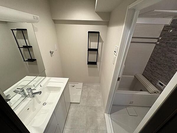 大きな鏡と一体型の洗面ボウルを備えた洗面化粧台の奥には洗濯機パンが設置されており、壁面には棚が配置された清潔感のある脱衣スペースです。隣接する浴室への動線もスムーズなレイアウトとなっています。