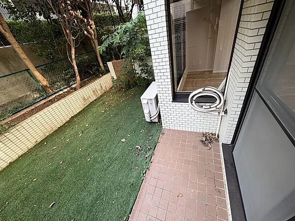敷地内には緑色の人工芝が敷かれた専用庭があり、室内の大きな窓に面したテラス部分はタイル敷きとなっております。敷地境界にはフェンスや植栽が配置され、建物の外壁は白いタイル貼りの仕様です。