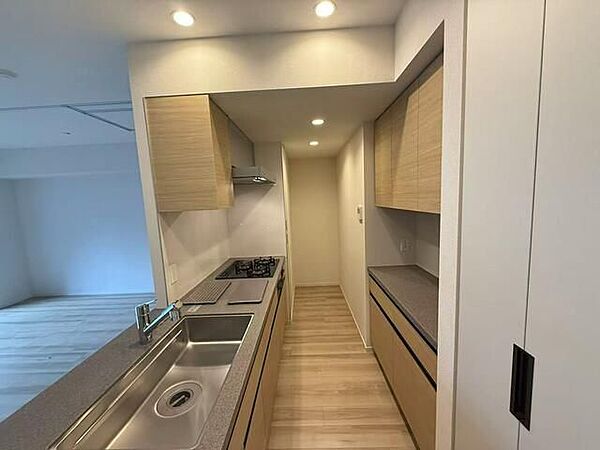 木目調の扉とグレーの天板をあわせた対面式システムキッチンには、3口ガスコンロやシンクが備わり、背面には上下の吊戸棚とカウンターが並ぶ収納豊富なカップボードが配置されています。