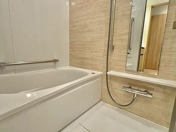 ベージュ系の木目調アクセントパネルを採用した浴室には、縦長の鏡やカウンター、高さ調節が可能なスライドバー付きシャワーのほか、浴槽横にも手すりが設置された仕様となっています。