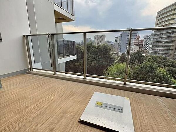 バルコニーには木目調シートの床材が施されており、手すりにはガラスパネルと可動式の物干し金物が備え付けられています。外部には緑豊かな樹木や周辺の建物を見渡せるロケーションが広がっています。