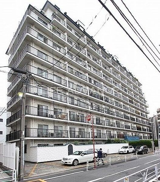 白を基調とした落ち着いたデザインの外観で、各住戸にバルコニーの手すりが整然と並ぶ集合住宅です。前面道路に面した敷地内には駐車場スペースや敷地を囲む白い塀が設置されています。