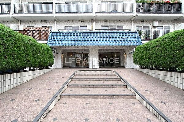 青い屋根が特徴的なエントランスアプローチには、中央の階段に加えて両脇にゆるやかなスロープが設けられており、アプローチの左右には整えられた生垣とタイル貼りの床面が広がっています。
