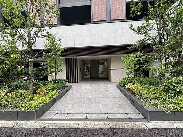 アプローチの両脇には青々とした植栽が整然と配されており、フラットなタイル敷きの通路を進むと、シックな縦格子デザインが施されたガラス扉のエントランスへとつながっています。