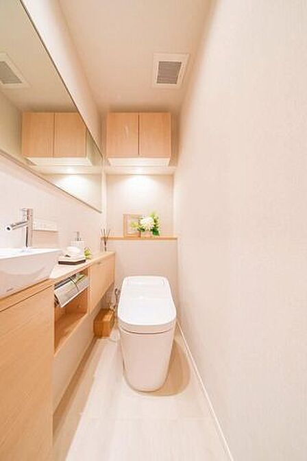 タンクレスタイプの便器を備えたトイレには、独立した手洗いボウルや大きな鏡が設置された木目調の手洗いカウンターに加え、上部に扉付きの吊戸棚収納も設けられています。