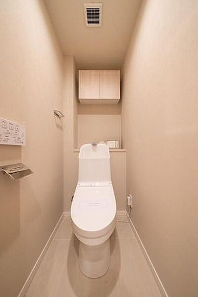 清潔感のある温水洗浄便座付きトイレには、壁面にリモコンやペーパーホルダーが設置されているほか、上部にはペーパーや清掃用品の保管に便利な木目調の吊戸棚収納が備え付けられています。