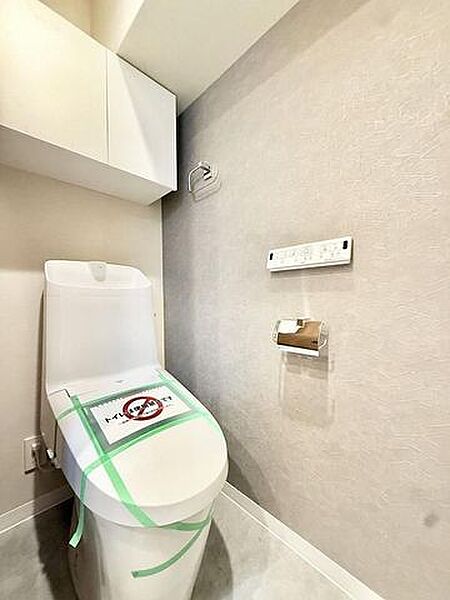 温水洗浄機能付きのトイレには、上部に備品を収納できる扉付きの吊戸棚が設置されているほか、壁面にはリモコンやタオル掛け、ペーパーホルダーが配置されており、グレー調のアクセントクロスが採用されています。