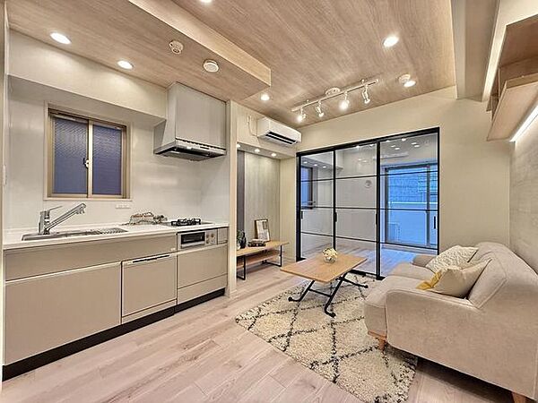 木目調の天井にダウンライトやライティングレールを備えたリビングで、奥には黒フレームのガラス製引き戸が設置されており、小窓やコンロの付いた壁付けのシステムキッチンも完備されています。
