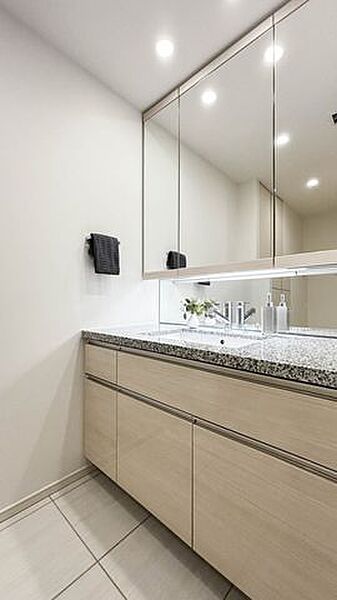 木目調のデザインと石目調のカウンターを採用した洗面化粧台には、大型の三面鏡や手元を照らす照明が付いており、足元には引き出し式の収納スペースや清潔感のあるタイル調のフロアが備わっています。