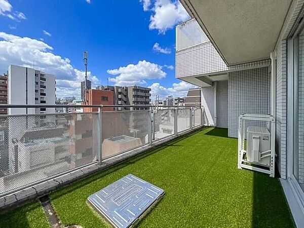 足元に鮮やかな緑色の人工芝が敷かれたバルコニーには、手すり部分に可動式の物干し金物が設置されており、避難ハッチやエアコン室外機用の架台といった設備も備えられています。