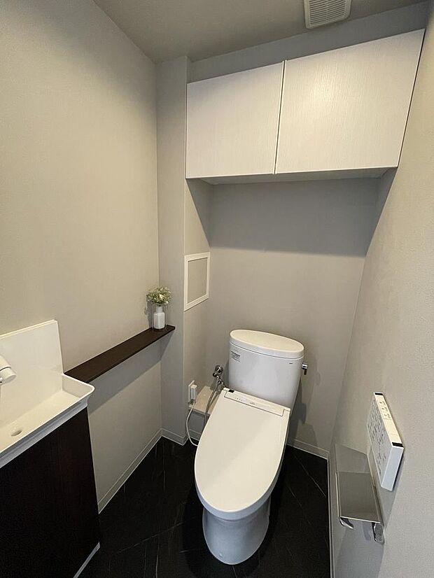 【トイレ】快適に使用できる手洗い場・温水洗浄便座仕様のトイレです。清潔感のある白い空間にダークカラーの床が、シックな雰囲気を演出しています。トイレ上部に吊戸棚が設けられており収納にも困りません。