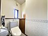 トイレ：小窓から光が差し込む、明るいトイレです。花柄のアクセントクロスが空間を彩ります。上部には棚があり、収納も可能です。