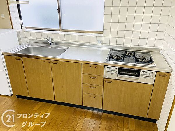 木目調のパネルが温かみを演出するキッチンは、三口コンロ採用で効率よくお料理を楽しめます。シンク前の窓から自然光が入り、明るく開放的な気分で家事に取り組める工夫が凝らされた設計です
