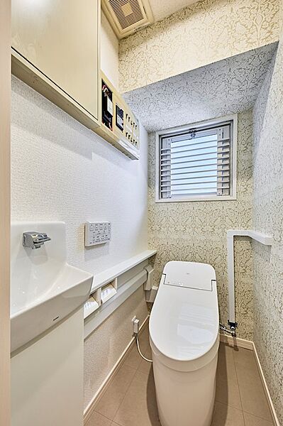 上品なデザインの壁紙が印象的なトイレ空間。手洗い器と窓があり、清潔感と開放感を両立しています。上部の収納でスッキリとした空間を保てます。