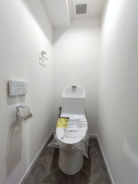 清潔感のある白い壁とヘリンボーン柄の床が調和した、落ち着いた雰囲気のトイレ空間。温水洗浄便座付きで、毎日を快適にお過ごしいただけます。