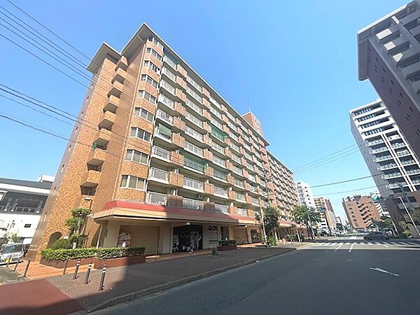 陽光が降り注ぐ中、整然とした外観のマンション。周辺には交通量の多い道路が走り、賑わいのある住環境が想像されます。