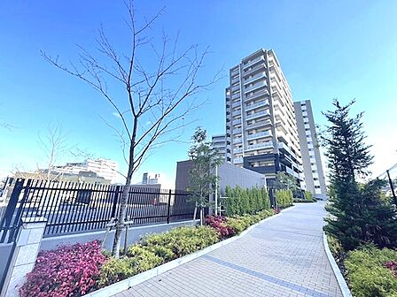晴れやかな青空が広がる現地写真です。手入れされた植栽と歩道が続き、住まいへの期待を高めます。高層マンションが視界に収まり、都市の暮らしを予感させます。