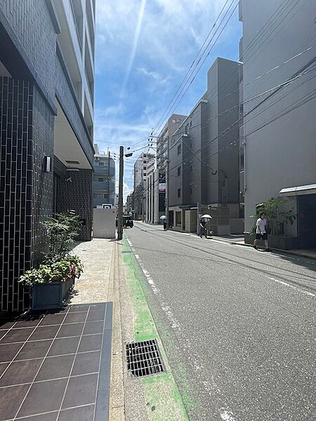 陽光が降り注ぐ、開放感のある道。周辺には商業施設やマンションが立ち並び、生活利便性の良い立地です。
