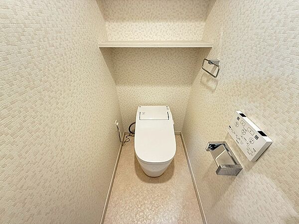 温水洗浄便座と便利な棚付きのトイレ空間。落ち着いた色合いのクロスが、リラックスできるひとときを演出します。