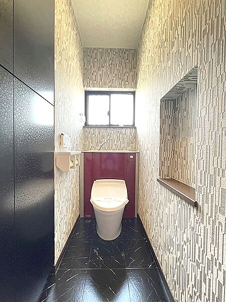 落ち着いた色合いの壁紙と黒い床が特徴のトイレ空間。窓から光が差し込み、明るく快適です。収納棚もあり、使い勝手の良さも魅力です。