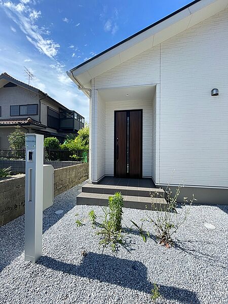 空の青と白い外壁が映える玄関。落ち着いたトーンの玄関ドアが、住まいの顔を穏やかに演出します。アプローチの砂利が明るく軽快な印象を与えます。