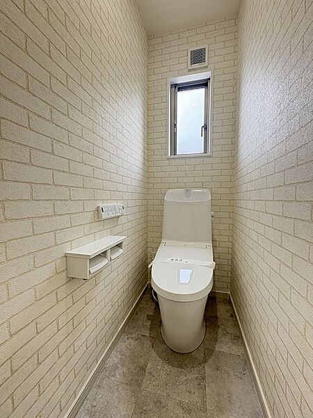 窓から自然光が差し込む明るいトイレ空間。レンガ調のクロスがおしゃれな印象を与えます。収納棚も備え付けられており、すっきりと整頓された空間を保てます。