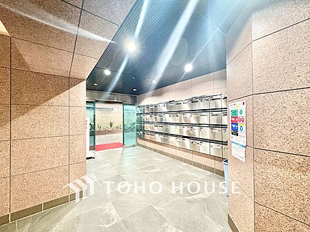 ガラス張りで明るい光が差し込む風除室には、高級感のある赤いマットが敷かれ、清潔に保たれています。 CSPのセキュリティ表示やAEDも設置されており、管理体制の良さが一目で分かります。