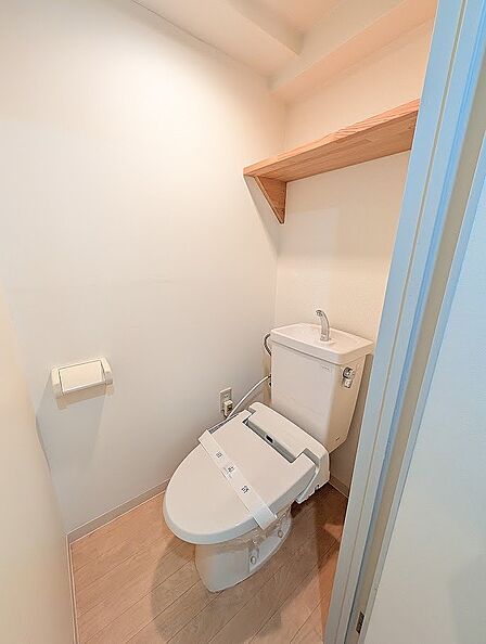 清潔感あふれる白を基調としたトイレです。上部にはトイレットペーパーや備品の保管に重宝する木製の固定棚を設置。手洗い付きタンクや温水洗浄便座を備えており、快適に利用できる空間になっています。